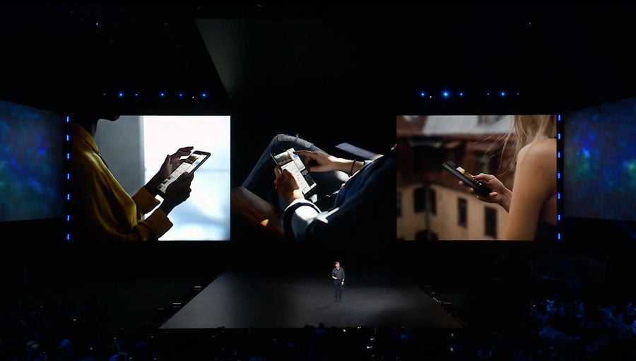 Evento de lanzamiento del Galaxy Fold. Se ve un hombre en un escenario oscuro con una pantalla gigante a sus espaldas, que muestra tres imágenes de uso del producto.