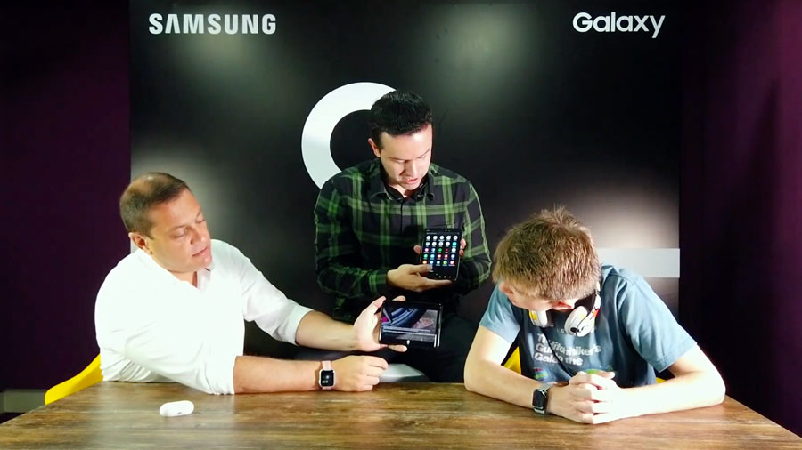 Tres hombres sentados frente a una mesa de madera y con un fondo de galaxy en sus espaldas, muestran la reproducción de video desde el dispositivo.